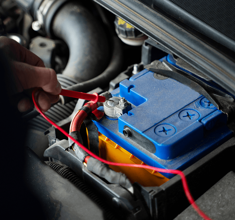 Close-up of a hand placing a red test lead on the positive terminal of a blue and yellow car battery in an engine bay